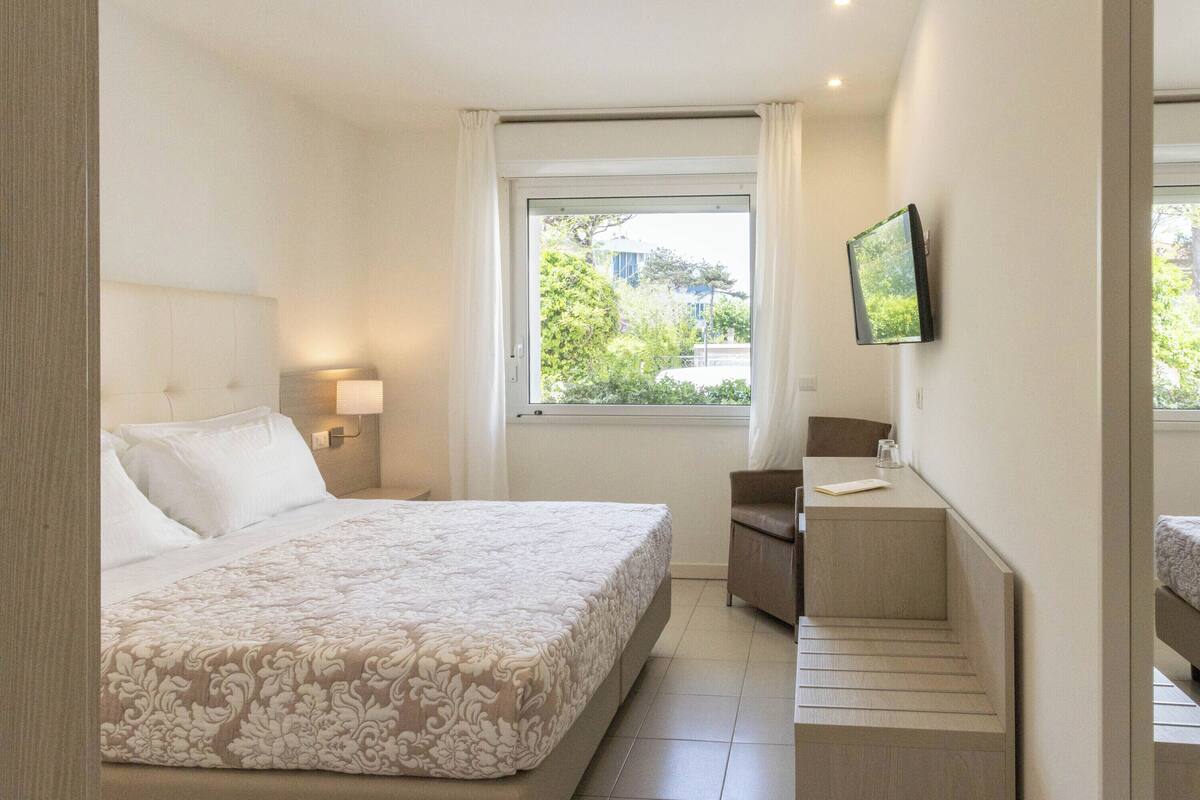 Habitación de hotel con cama, mesita de noche, televisor y ventana con vistas a la vegetación.