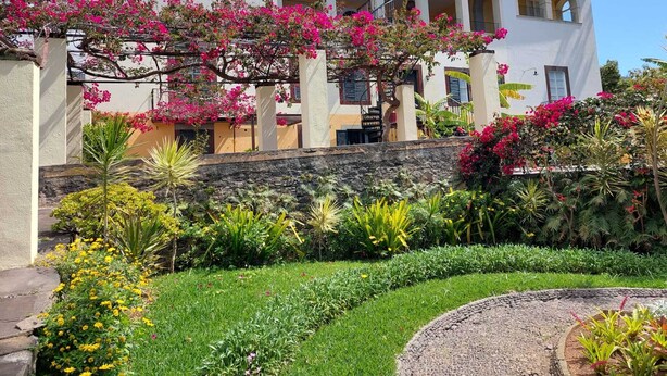 A white building with arched windows and a balcony, surrounded by a well-maintained garden with various plants and flowers. - Cozy Studio in a fabulous Quinta with pool and sea view! (Funchal)