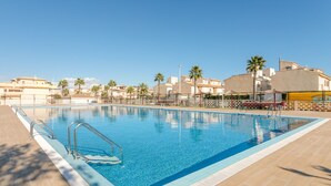 Una piscina con plataforma de baldosas, palmeras y edificios al fondo.