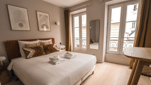 A neatly made bed with white linens and two towels, a bedside table with a lamp, and a window with a view of buildings. - Appartements rue Amélie (Paris)
