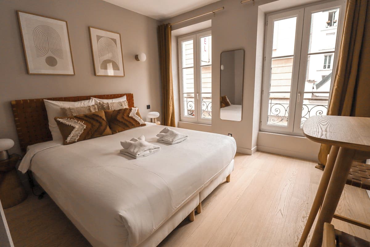 Un lit bien fait, avec du linge de lit blanc et deux serviettes, une table de chevet avec une lampe, et une fenêtre donnant sur des immeubles.