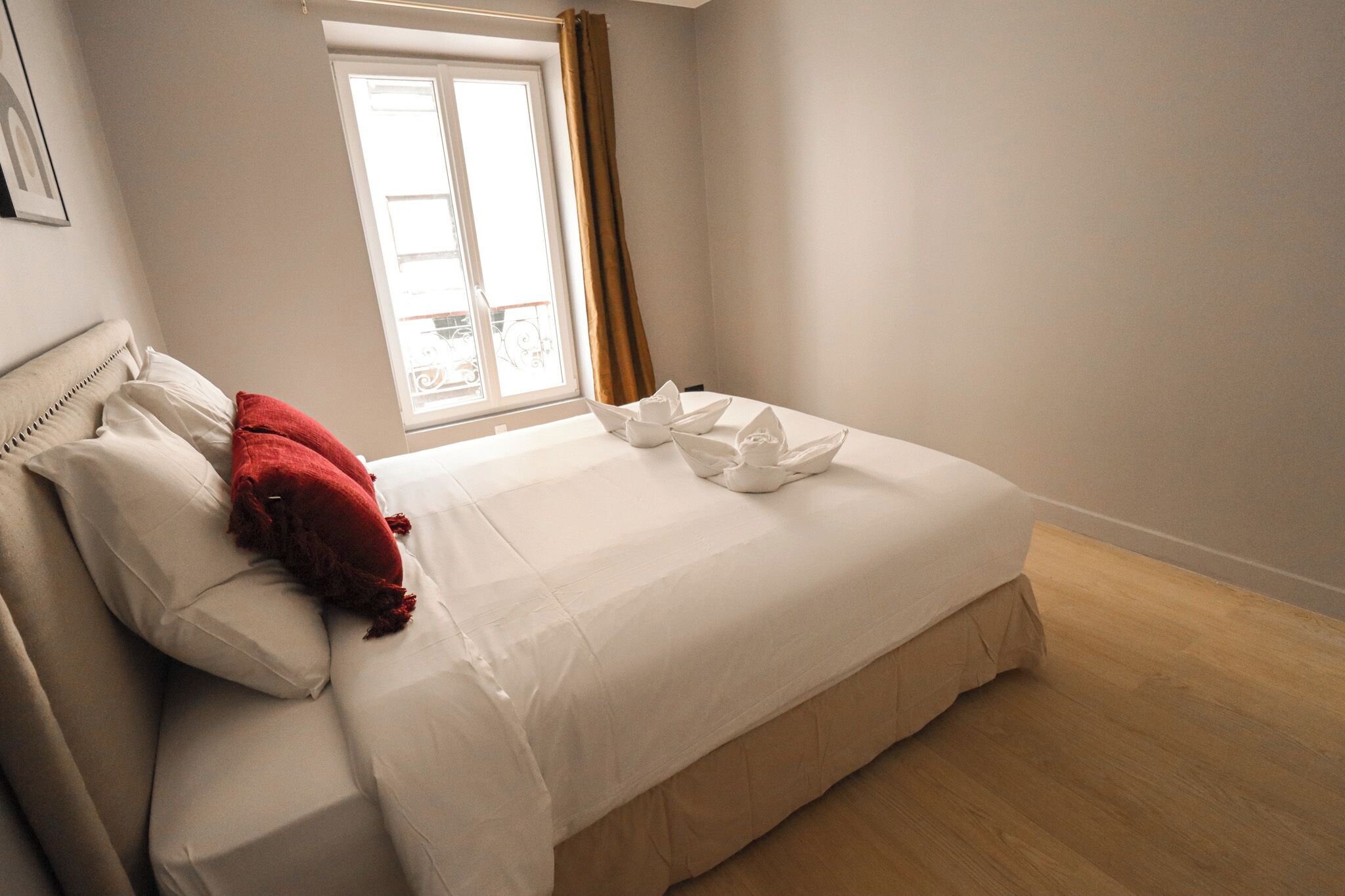 Ein ordentlich bezogenes Bett mit weißen Leinen und einem roten Kissen, ein Fenster mit Vorhängen und ein Holzboden.