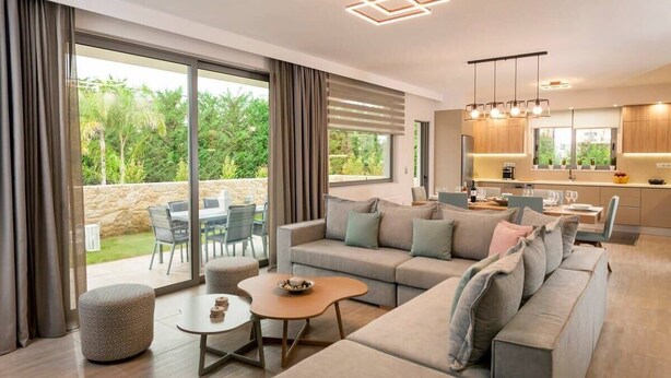 Una sala moderna con sofá, reposapiés y mesa de centro, un comedor con sillas y una cocina con armarios de madera e iluminación colgante.
