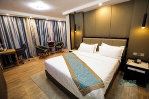 A hotel room with a large bed, a desk with a chair, a small table with a vase, and a nightstand with a phone.