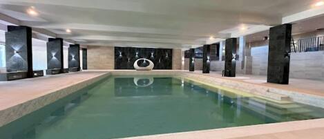 Ein großes Hallenbad mit einem zentralen, kreisförmigen Element, umgeben von dunklen Säulen und einem gefliesten Boden.