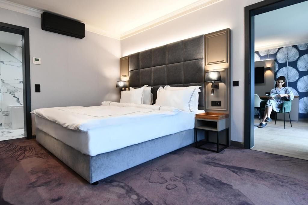 A modern hotel room with a large bed, bedside lamps, a small table, and a view of a dining area.