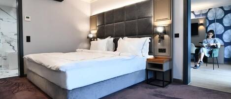A modern hotel room with a large bed, bedside lamps, a small table, and a view of a dining area.
