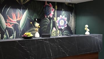 En receptionsdisk med en väggmålning med blommotiv, en skål med frukt och en vas.