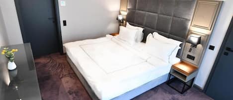A hotel room with a large bed, bedside tables with lamps, a desk with a vase of flowers, and a door.