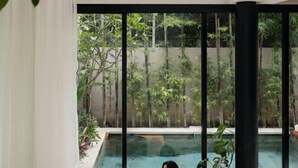 A modern indoor pool area with clear glass walls, surrounded by lush greenery and bamboo plants.