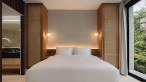 A modern hotel room with a large bed, wooden headboard, and a view of greenery through the window.