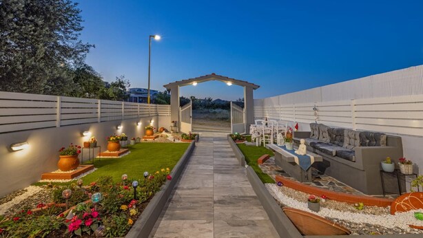 A well-lit outdoor patio area with a pathway leading to a covered seating area, surrounded by decorative plants and a white fence. - Ranias House Faliraki (Rhodes)
