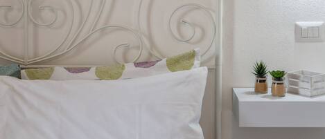 Un lit avec une literie blanche et des coussins décoratifs, une tête de lit blanche au design incurvé et une table de chevet avec une petite plante et un pot.