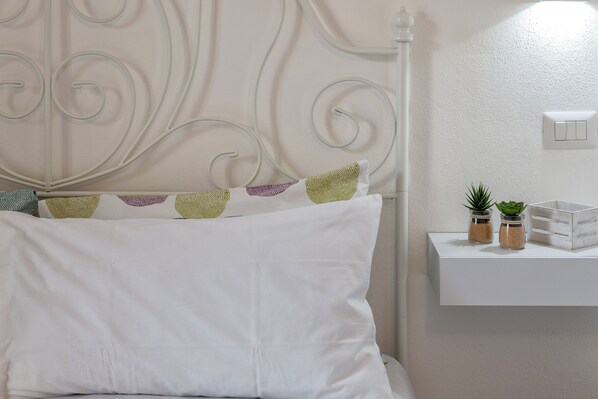 A bed with white bedding and decorative pillows, a white headboard with a curved design, and a bedside table with a small plant and a jar.