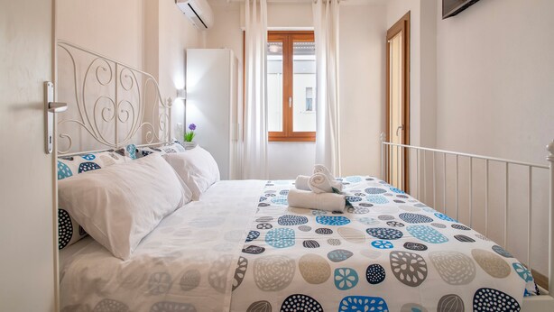 Quarto com cama, janela com cortinas, TV fixada na parede e uma porta.