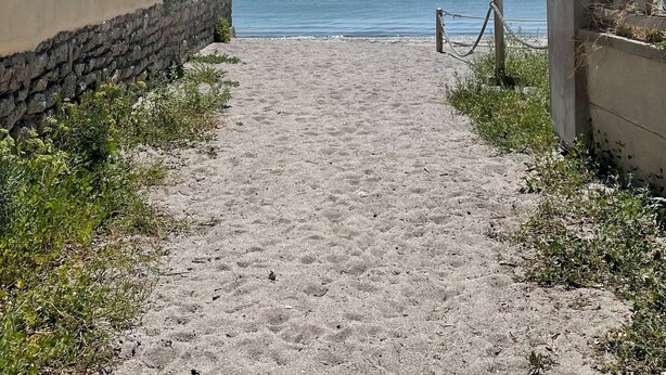 Un camino de arena que lleva al mar, con un muro de piedra a un lado y una reja metálica al otro.