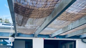 Un moderno espacio lounge al aire libre con una silla blanca acolchada, un ventanal de vidrio que da a una piscina y un porche cubierto con techo de fibras naturales.
