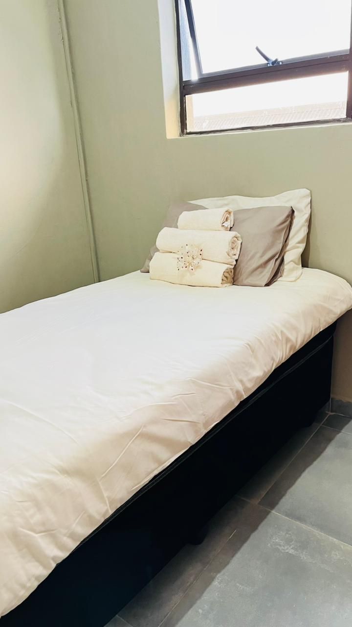 Una cama individual con una colcha y almohadas color beige, una ventana arriba y suelo de baldosas.