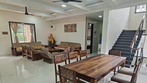 A spacious living area with a wooden dining table, sofas, and chairs. There are ceiling fans and a staircase in the background. - Mansarovar Bungalows (Lonavala)