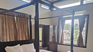 A four-poster bed with a dark frame, a white mattress, and two pillows. There is a wooden wardrobe to the right of the bed and a window with curtains to the left. - Mansarovar Bungalows (Lonavala)
