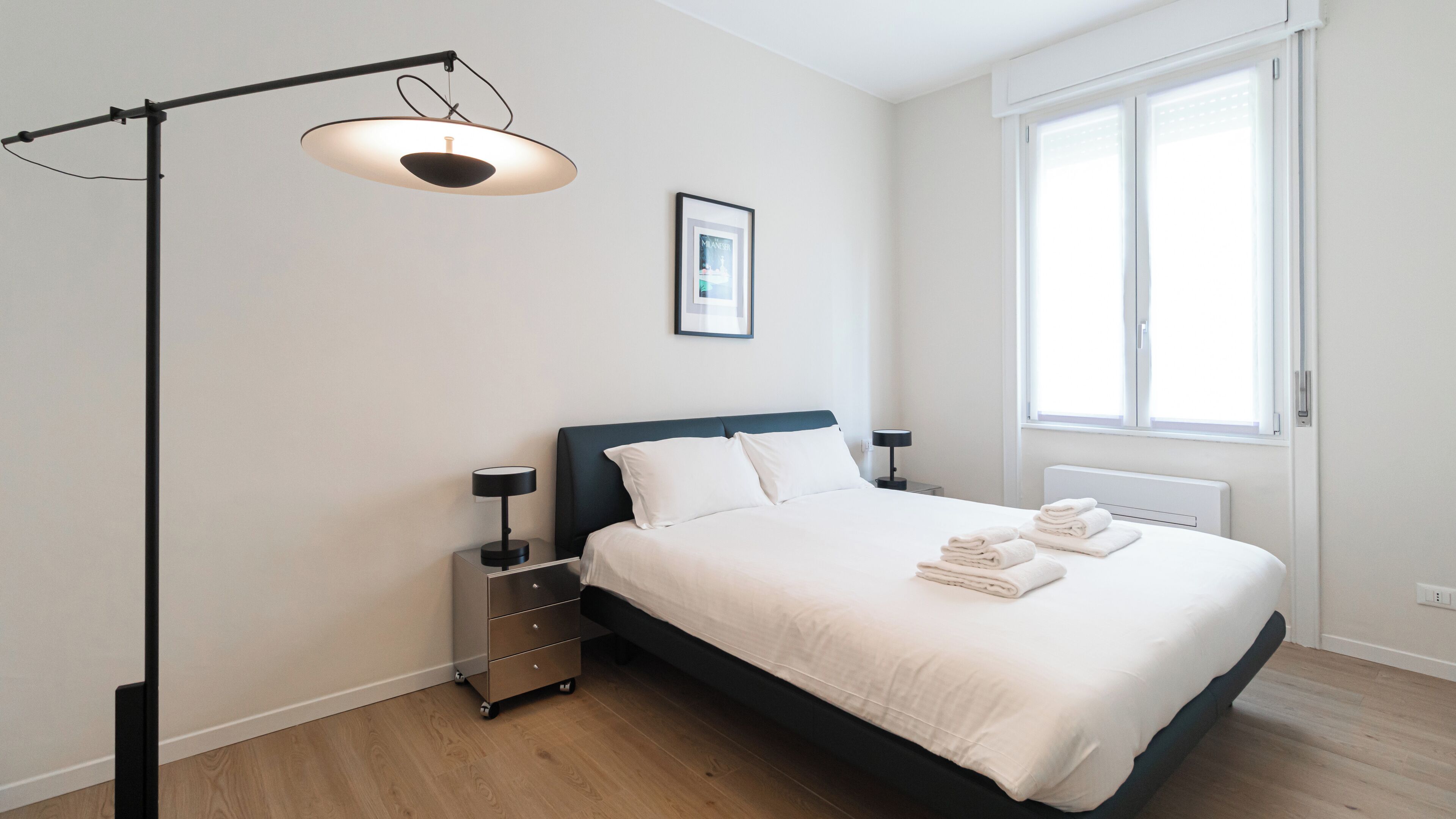 A modern bedroom with a bed, bedside tables, a floor lamp, a window, and a framed picture on the wall.