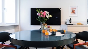 A modern dining area with a round glass table, chairs, a vase of flowers, breakfast items, and a flat-screen TV on the wall. - Italianway Easy - Sebastiano Veniero 27 C - Bilo (Milan)