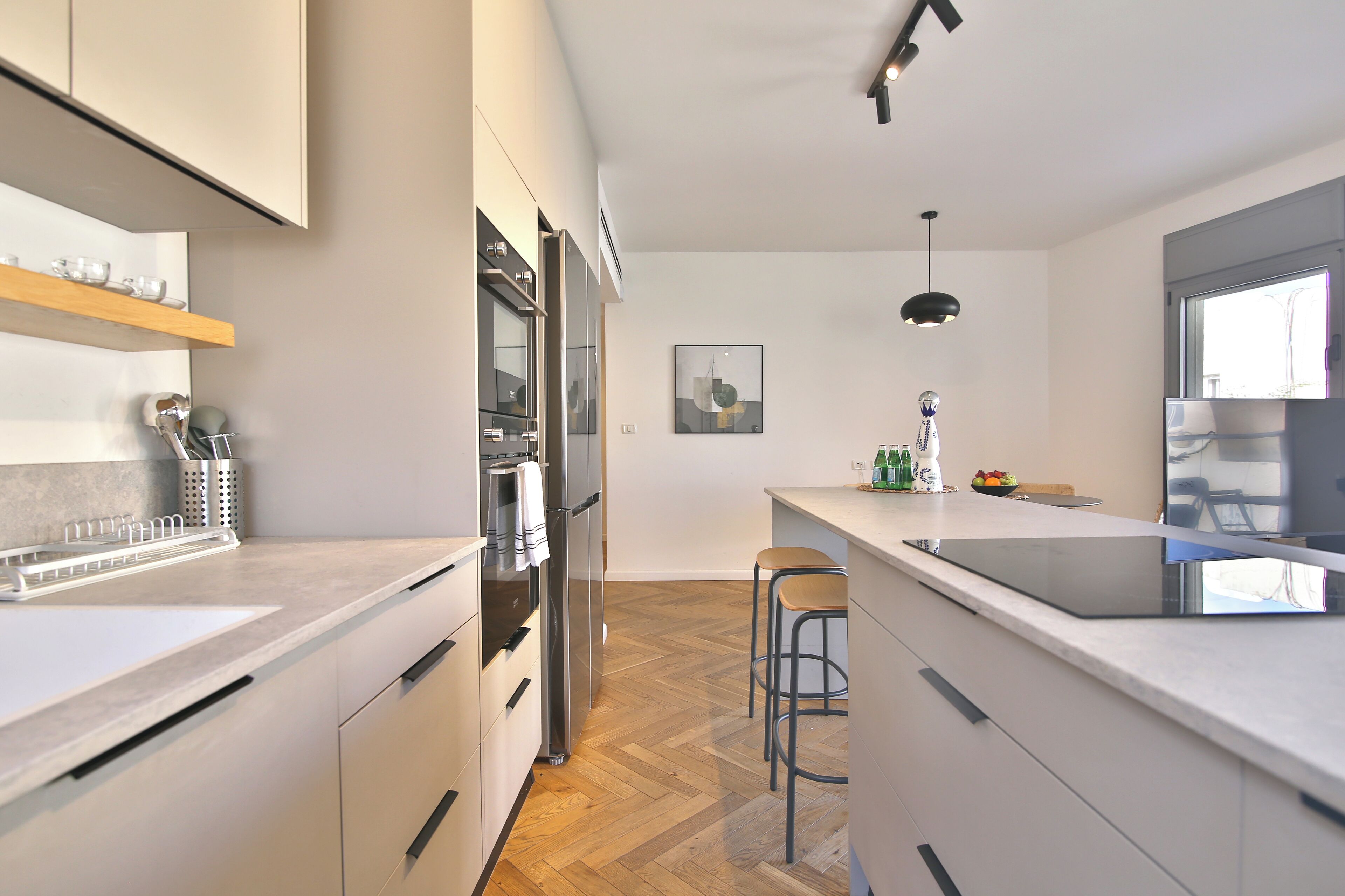 A modern kitchen with white cabinets, a wooden herringbone floor, a stainless steel refrigerator, and a bar area with stools.