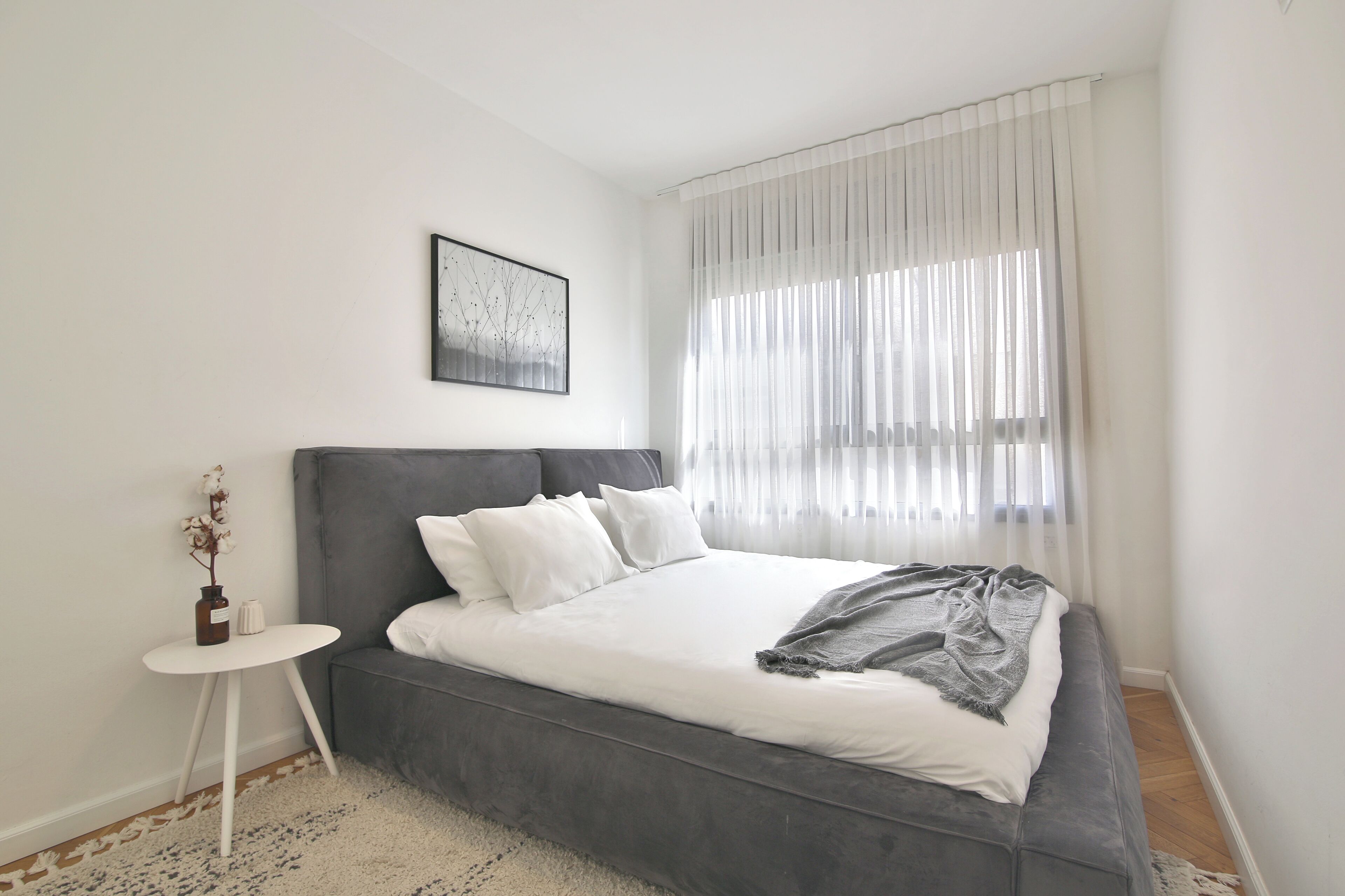 Een moderne slaapkamer met een grijze slaapbank, wit beddengoed, een klein wit nachtkastje en een lijst met een afbeelding aan de muur.