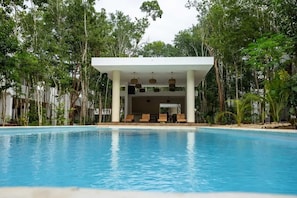 Una piscina con sillas de descanso y un área de estar cubierta, rodeada de árboles.