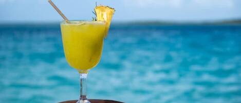 A glass of yellow drink with a straw and a pineapple slice on a wooden stump by the ocean.