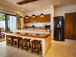 A modern kitchen with wooden cabinets, a central island with stools, pendant lighting, and a black refrigerator.