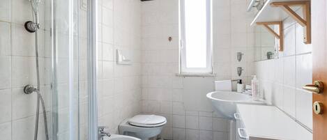 A bathroom with a glass shower enclosure, two toilets, a bidet, a sink, and a window providing natural light.