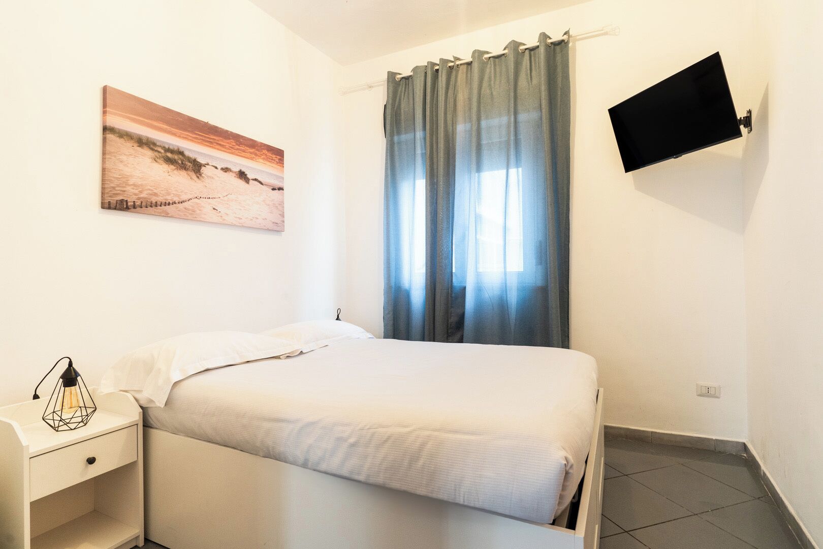 Un dormitorio con cama, mesita de noche, un cuadro, televisor montado en la pared y ventana con cortinas.