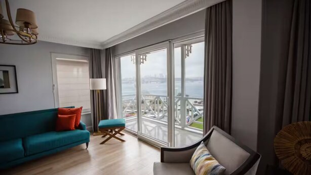 Uma sala de estar moderna com um sofá verde-água, um banquinho pequeno e uma cadeira com uma almofada colorida. Uma janela grande oferece vista para uma ponte e para a água.