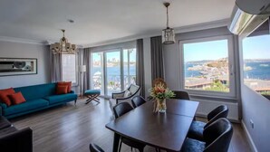 Uma sala de estar moderna com sofá, mesa de jantar e cadeiras. Há uma janela grande com vista para a cidade e o mar.