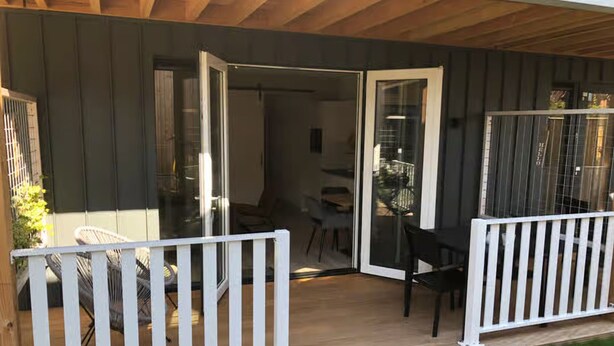 Un espacio al aire libre techado con una terraza de madera, barandas blancas y una puerta de vidrio que da a un ambiente interior con una mesa y sillas.