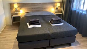 Una habitación de hotel moderna con una cama grande, dos mesitas de noche con lámparas y vistas al exterior a través de una ventana con cortinas.