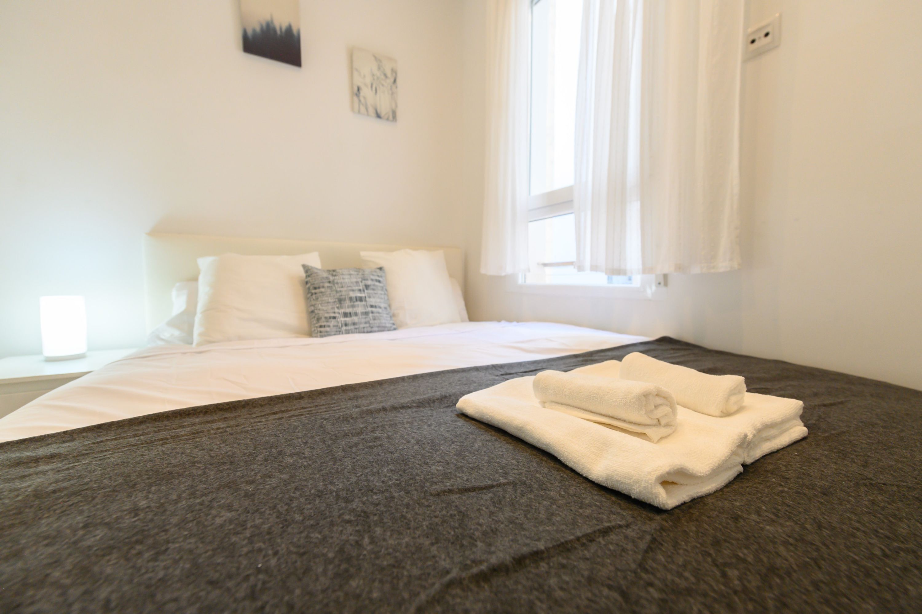A neatly made bed with a grey bedspread and white pillows, a folded towel on the bed, and a window with sheer curtains.