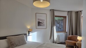 A bedroom with a bed, a chair, a window with curtains, a picture on the wall, and a lamp on a nightstand. - 3-Room Apartment in Valmorel - Sleeps 6, Parking (Valmorel)