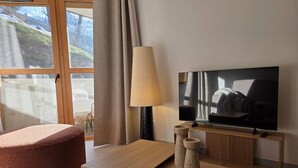 A modern hotel room with a large window, a flat-screen TV, a wooden coffee table, a red cushioned chair, and a beige lamp. - 3-Room Apartment in Valmorel - Sleeps 6, Parking (Valmorel)