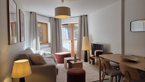 A modern living room with a sofa, a coffee table, a dining table with chairs, a TV, and a large window with curtains. - 3-Room Apartment in Valmorel - Sleeps 6, Parking (Valmorel)