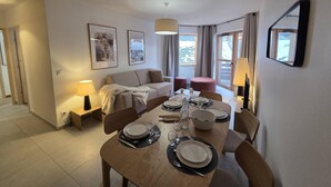 Une chambre d’hôtel moderne avec un coin repas aménagé pour quatre personnes, un canapé, une petite table et une fenêtre avec des rideaux.