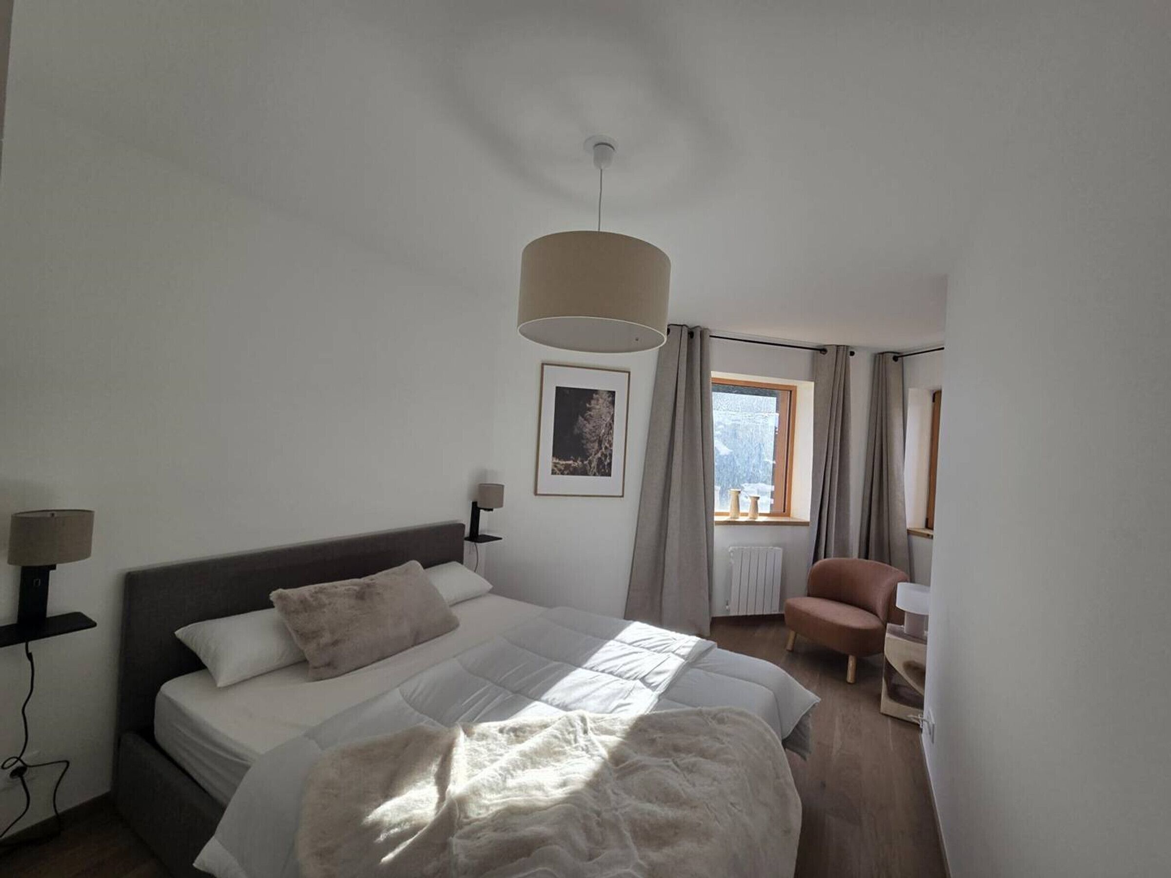 Una camera da letto con un letto, lampade sui comodini, una finestra con tende, una sedia e un termosifone.