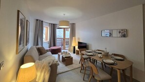 Un salon moderne avec un canapé, une table à manger pour deux personnes et une petite table basse. Il y a une fenêtre avec des rideaux, une lampe et un miroir au mur.