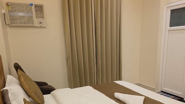 A hotel room with a bed, pillows, a brown blanket, a white towel, a window with curtains, and an air conditioning unit on the wall. - Sarvagya Inn (Varanasi)