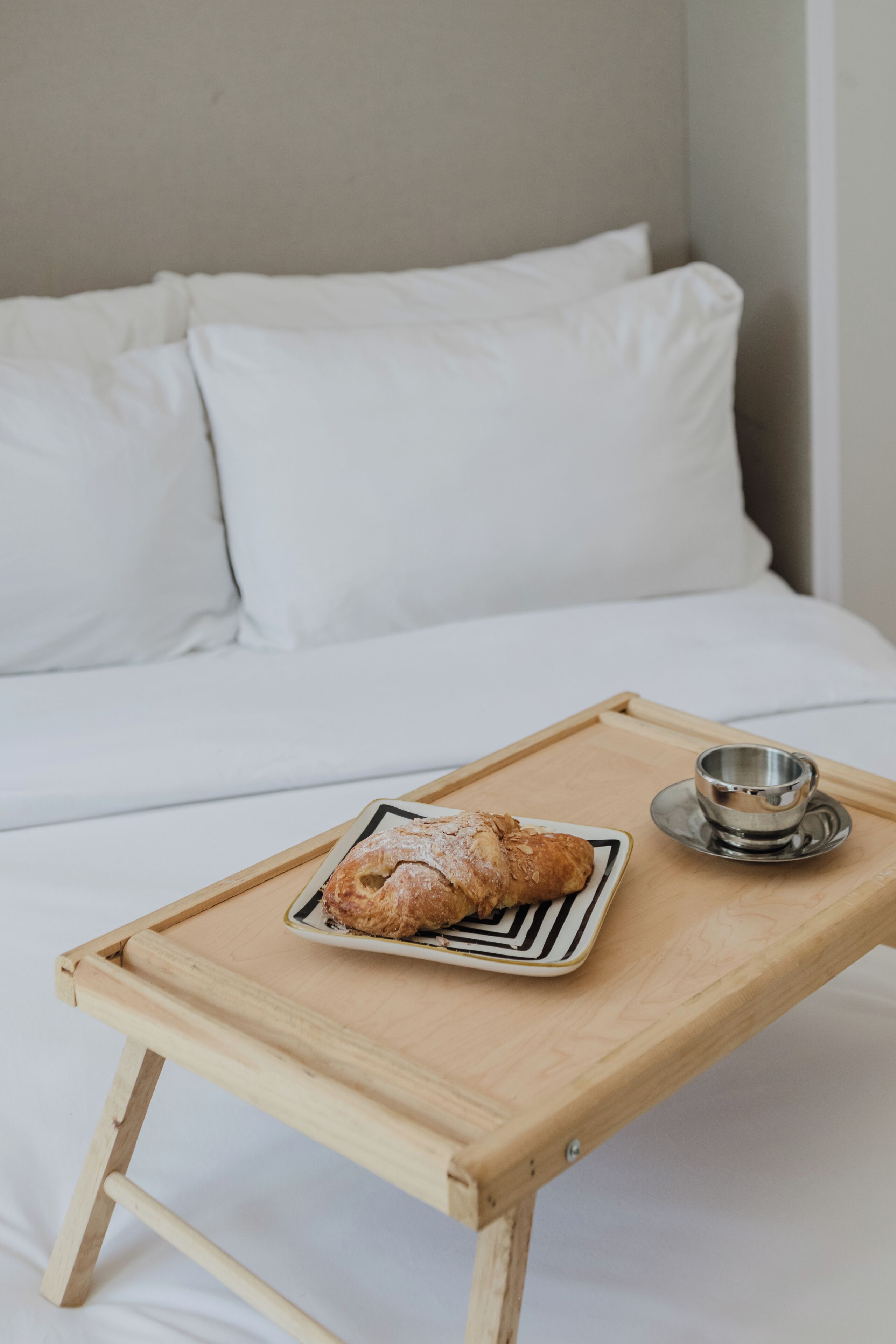 Uma bandeja de madeira com um croissant e uma xícara sobre uma cama com roupa de cama branca.