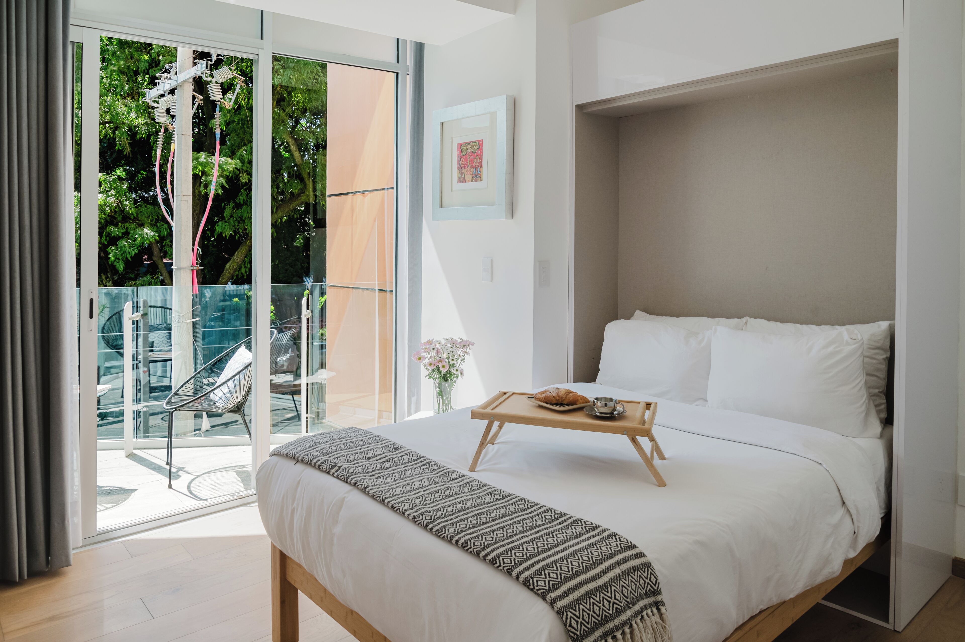 A modern hotel room with a large bed, a wooden bedside table with a tray, and a view of a balcony with outdoor furniture.