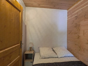 Ein kleines Zimmer mit einem Einzelbett, einem Holzschrank, einem Nachttisch mit Lampe und einer schlichten weißen Wand.