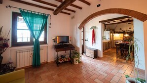 Un salon confortable avec un téléviseur, une petite table et une fenêtre ornée de rideaux verts. On peut apercevoir une cuisine à travers une arche.