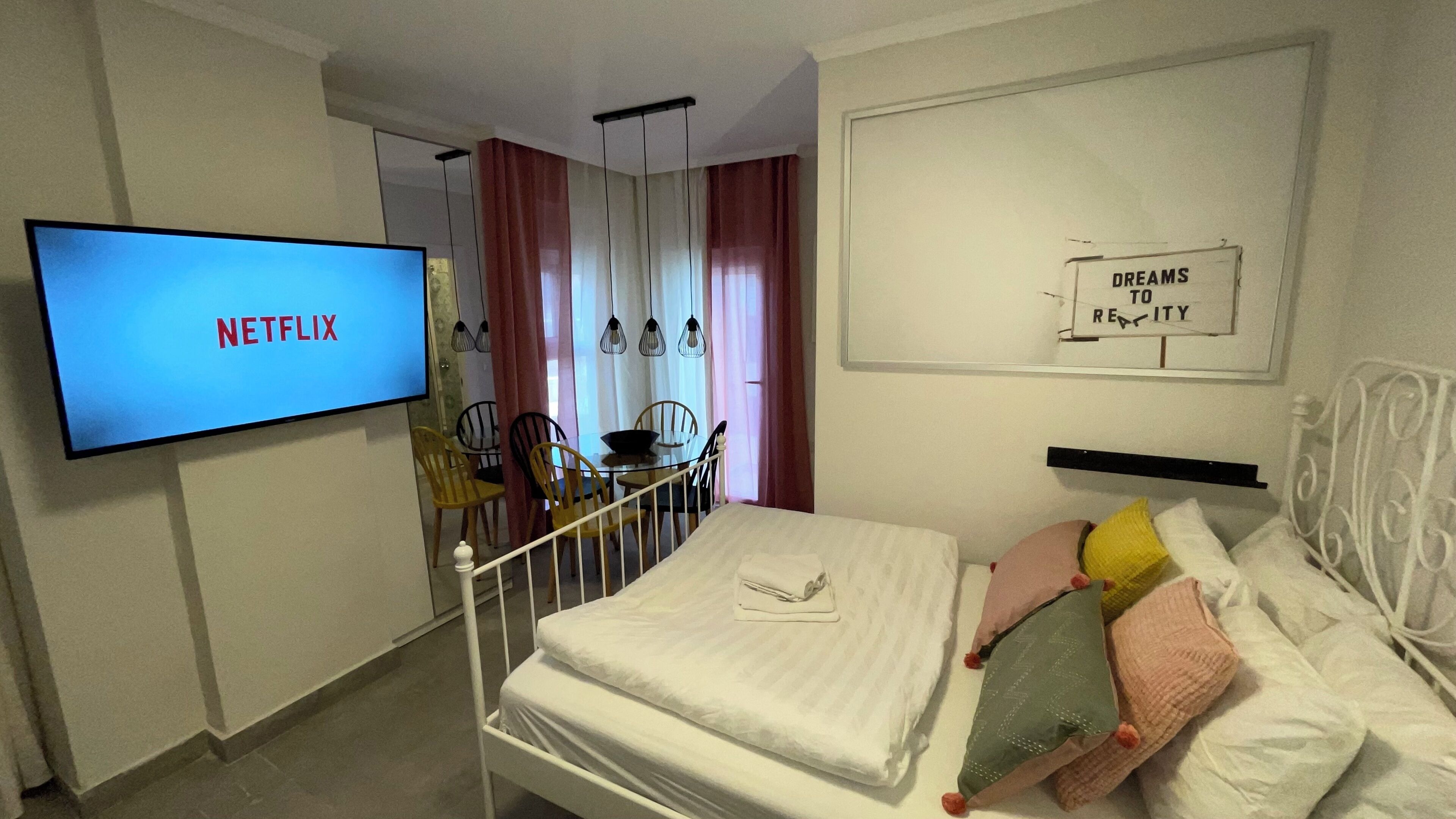 Ein Hotelzimmer mit einem Bett, einem Fernseher, der 'NETFLIX' anzeigt, einem Essbereich mit Stühlen und einem Schild an der Wand mit der Aufschrift 'DREAMS TO READ'.